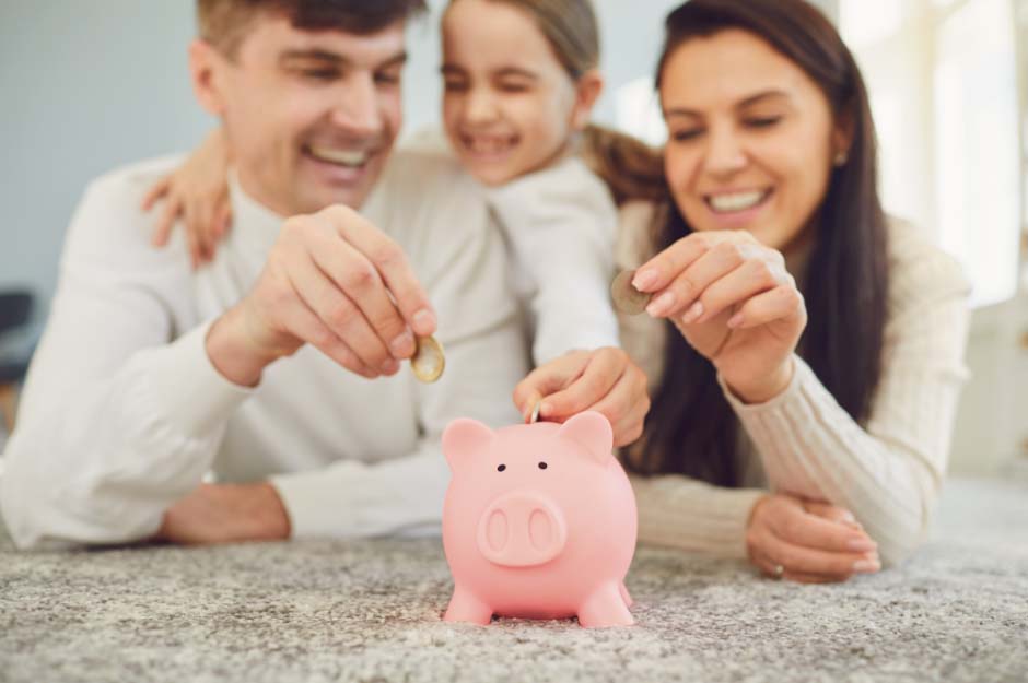 A younger couple with small child put pound coins into a traditional looking piggy bank. the image is used to symbolise Remortgages offered by Whitewood Mortgages.